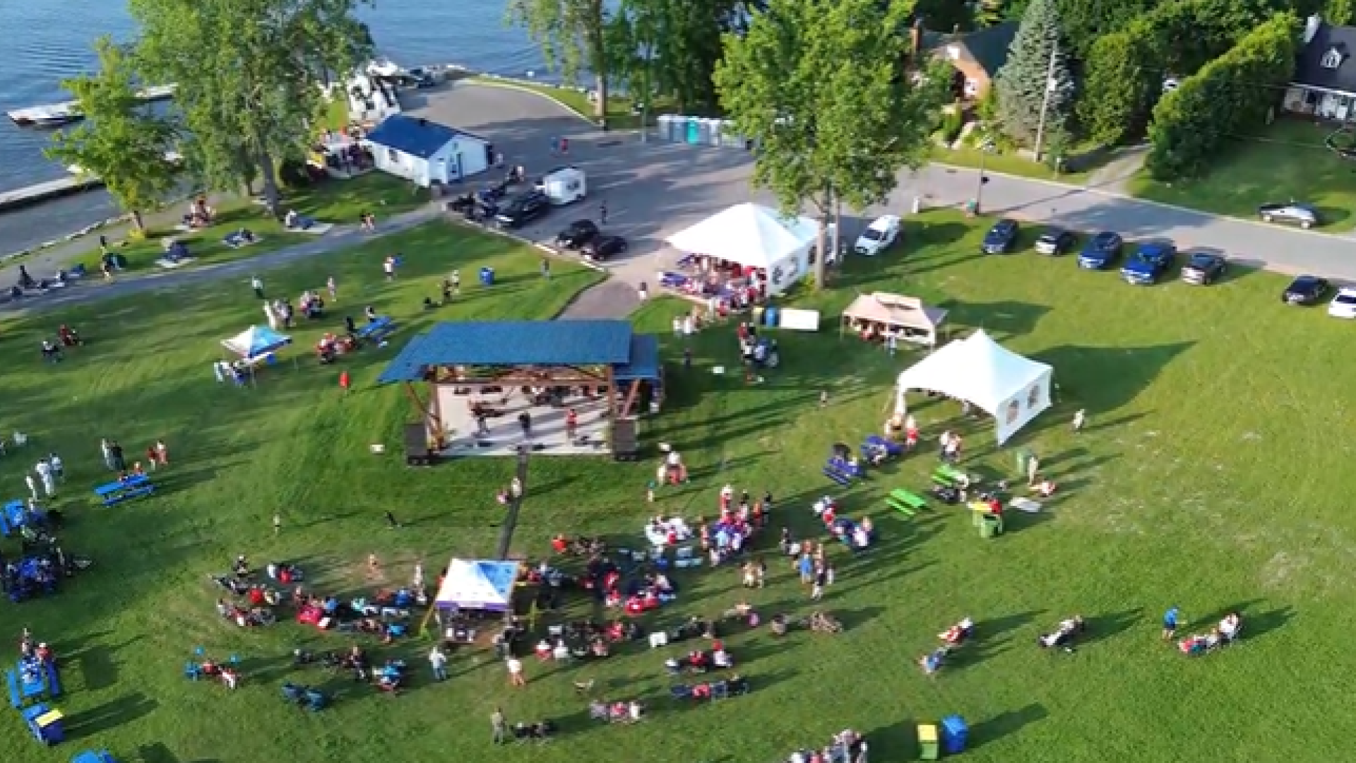 Aerial view of a lakeside park event with tents and people on a green lawn.