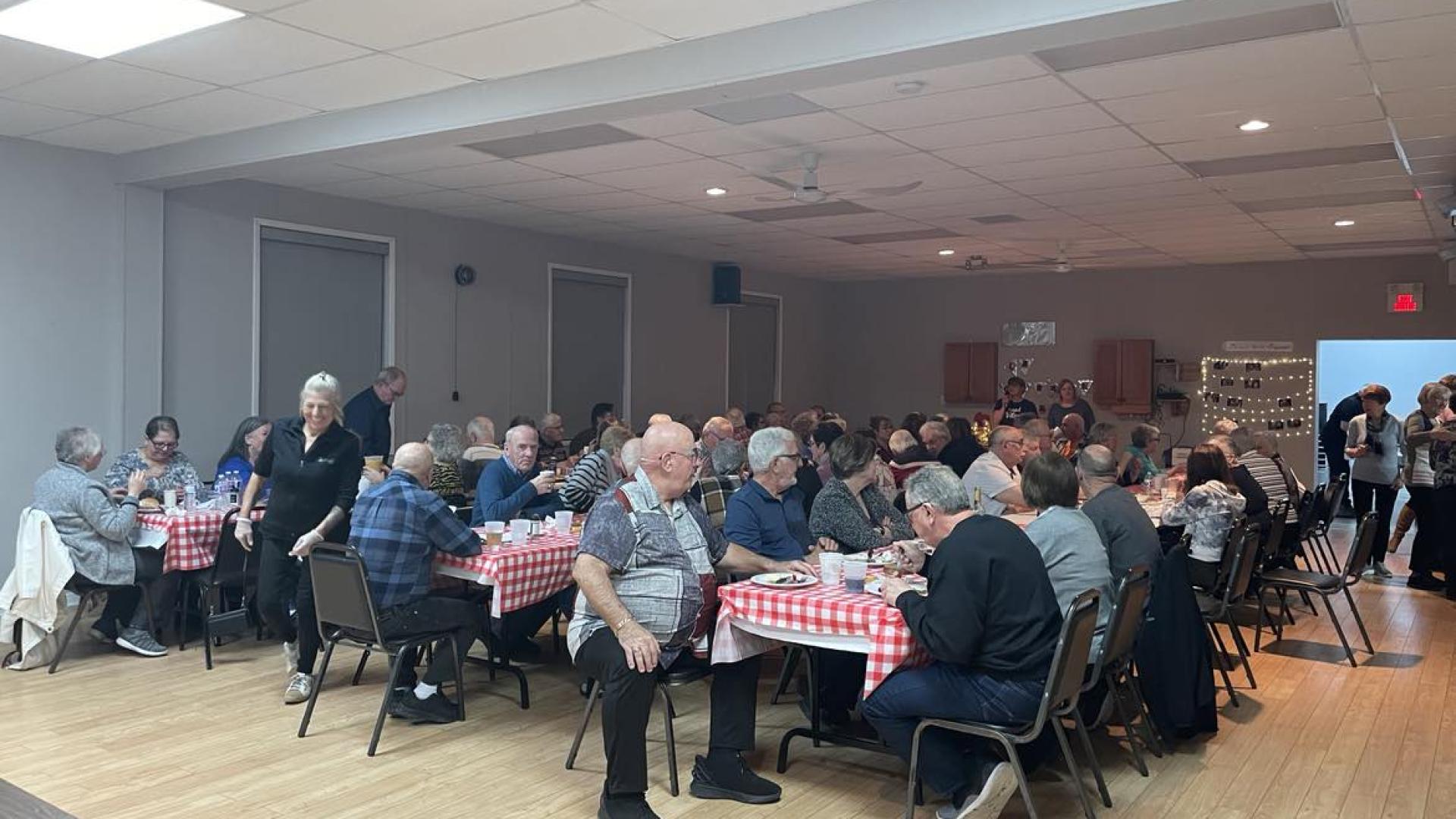 People seated at tables in a community hall with red checkered tablecloths.