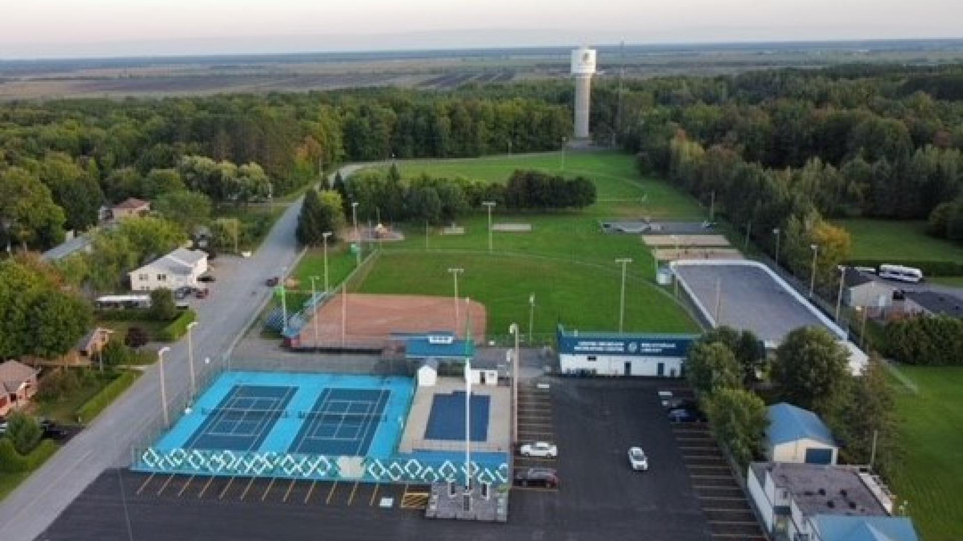 Aerial view of a sports complex with pools, tennis courts, and fields surrounded by trees.