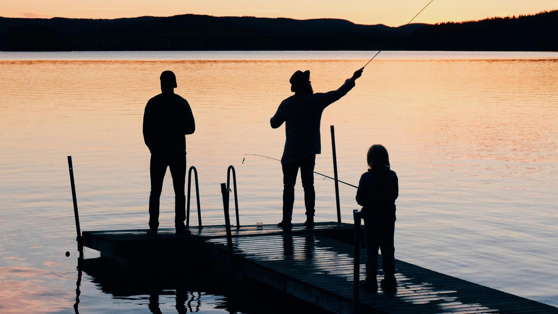 Three people fishing on a dock at sunset, silhouetted against the water.