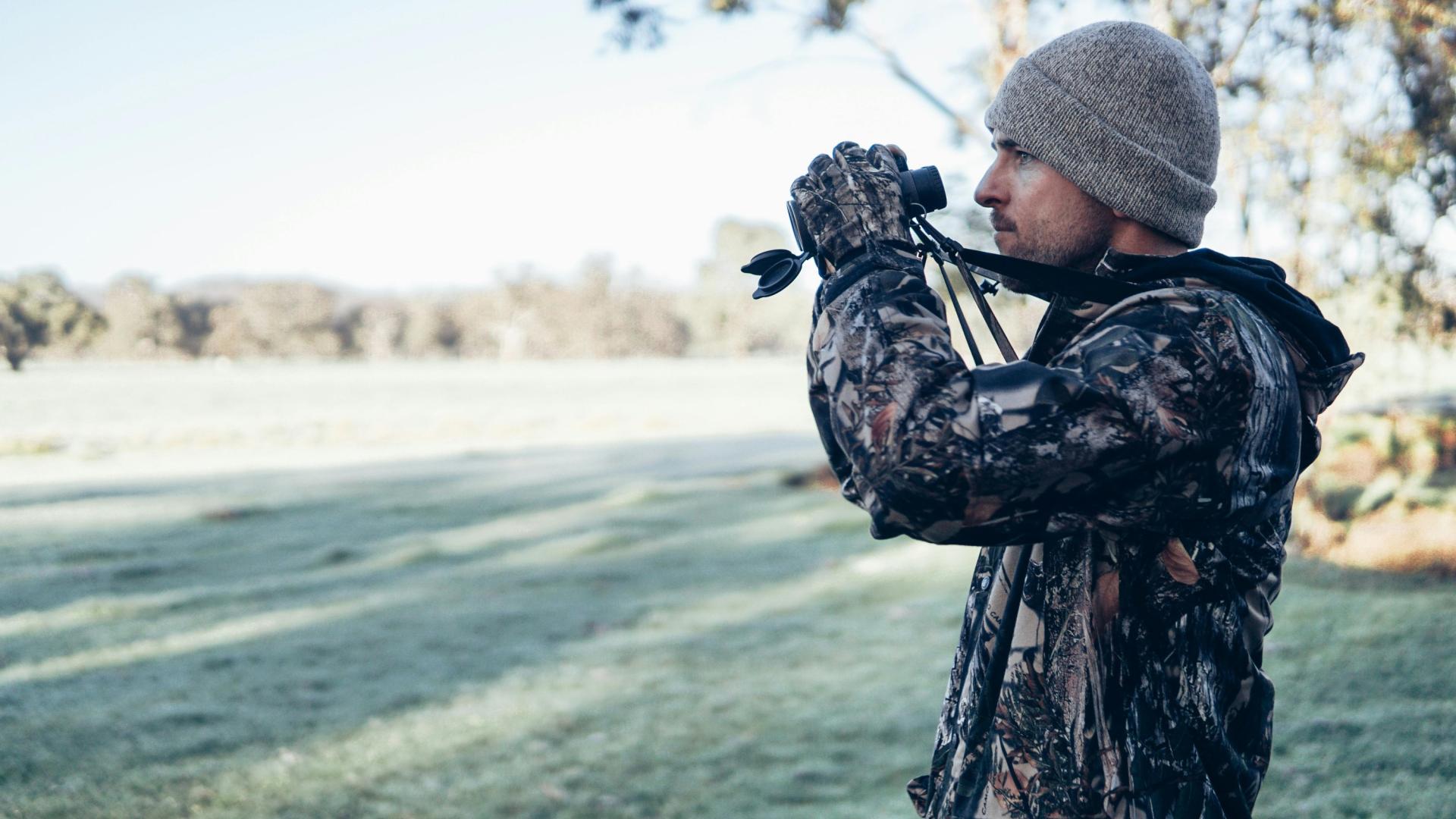 Man in camo gear using binoculars on a frosty field.