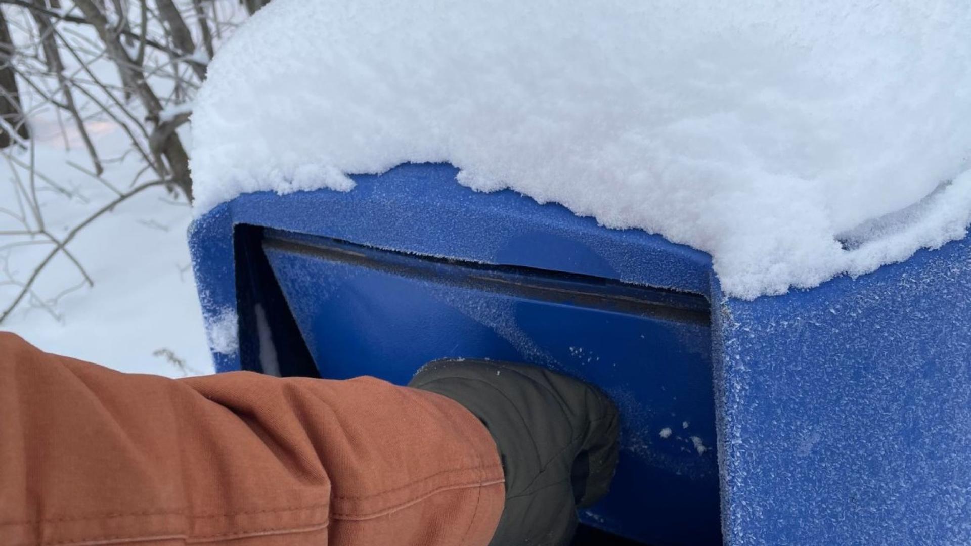 Hand in a glove putting something into a snow-covered blue trash can in a snowy forest.