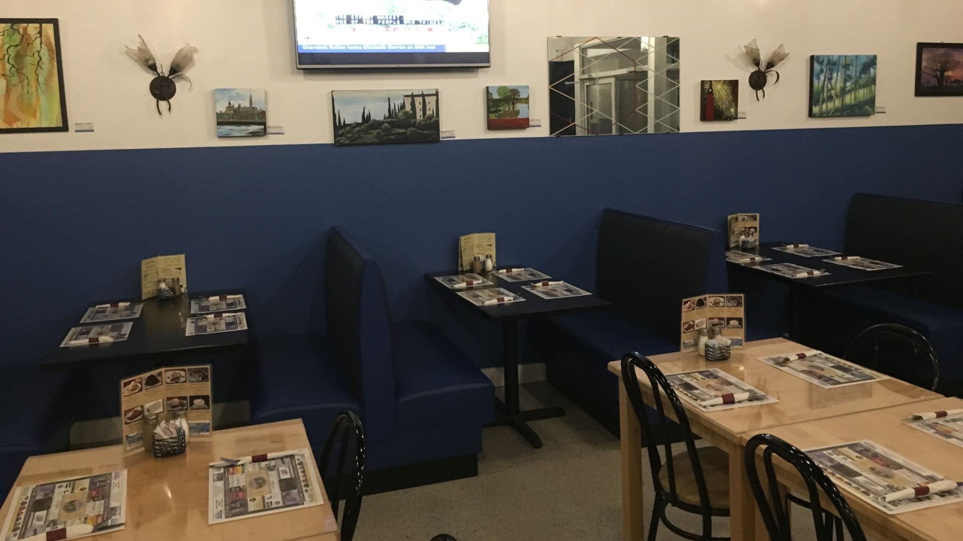 Restaurant interior with blue booths, tables set with menus and placemats, artwork on walls.