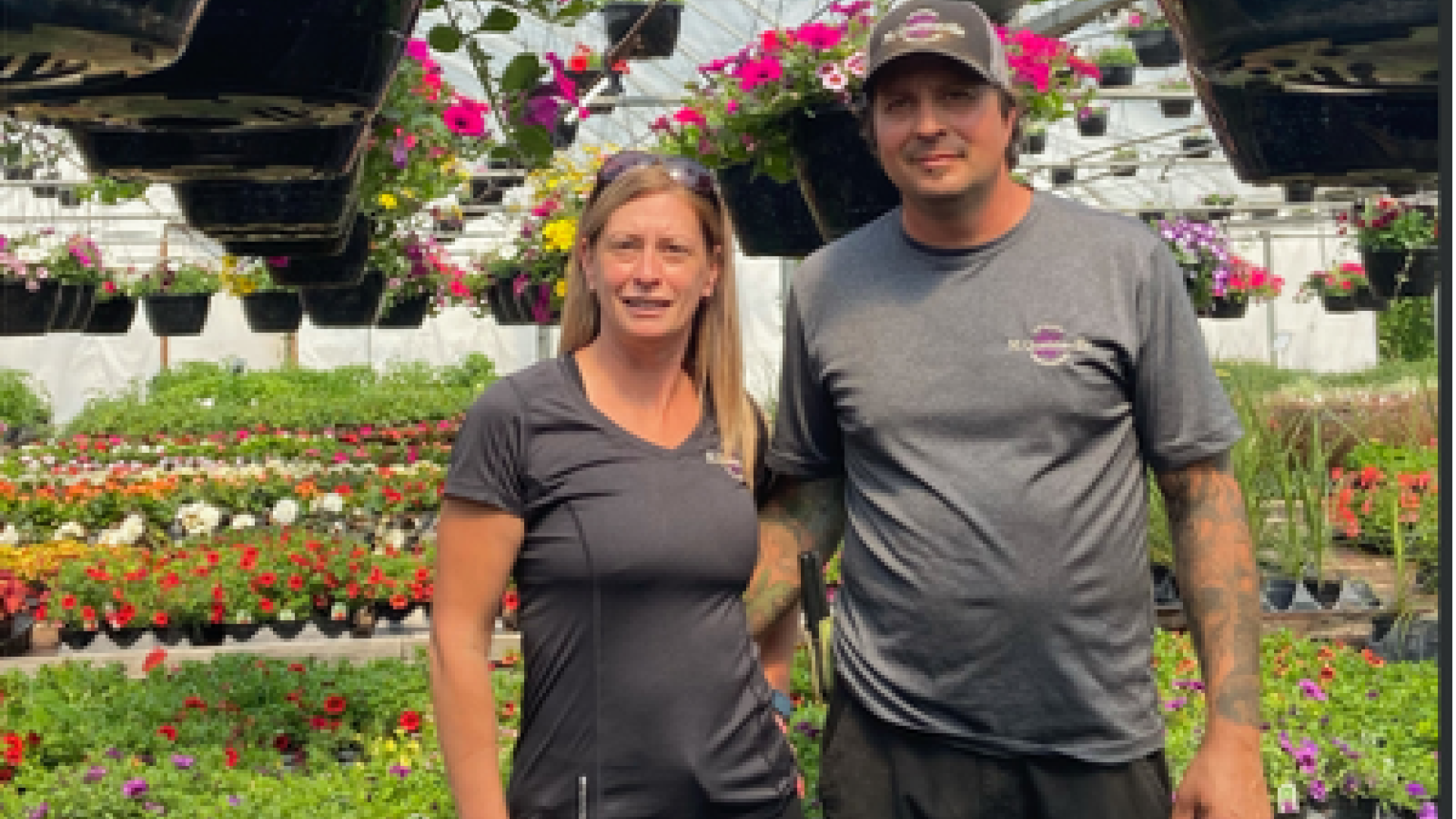 Two people stand in a colorful garden greenhouse, smiling.