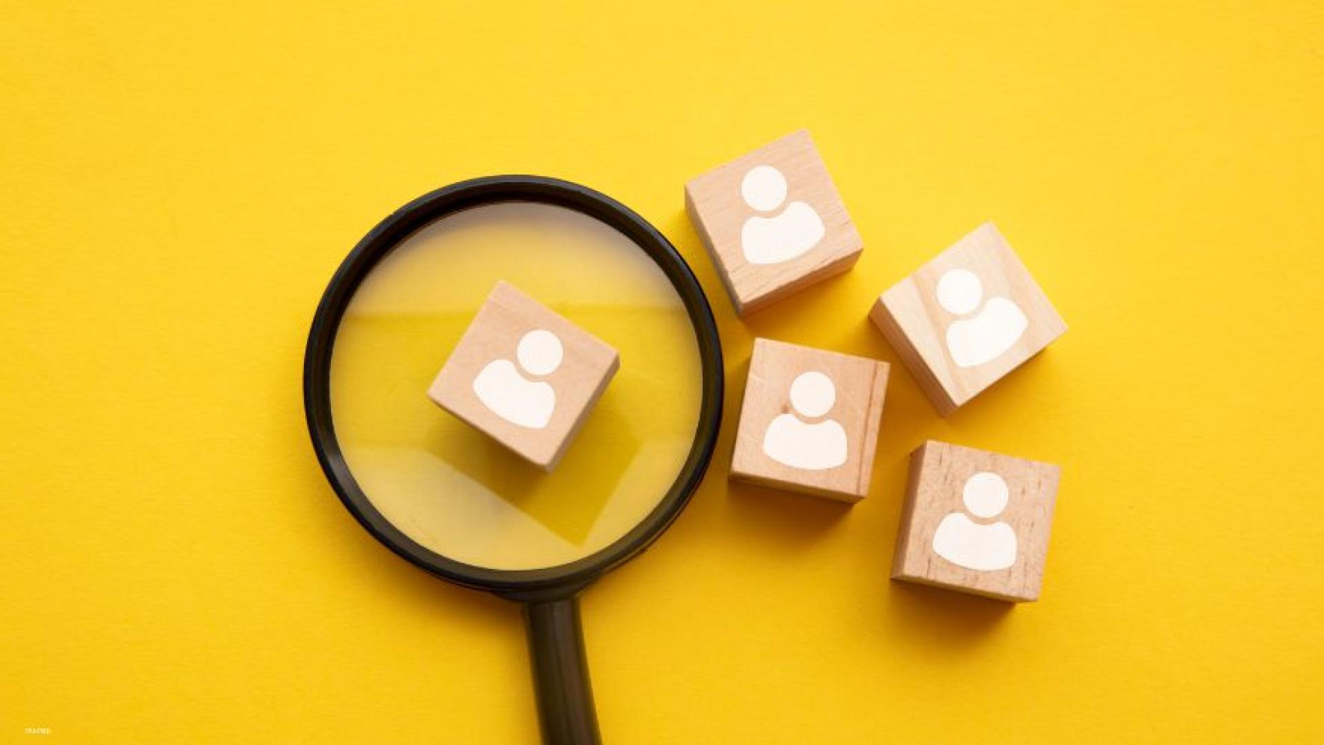A magnifying glass highlighting small wooden cubes with people icons on them, symbolizing that the organization is searching for employees