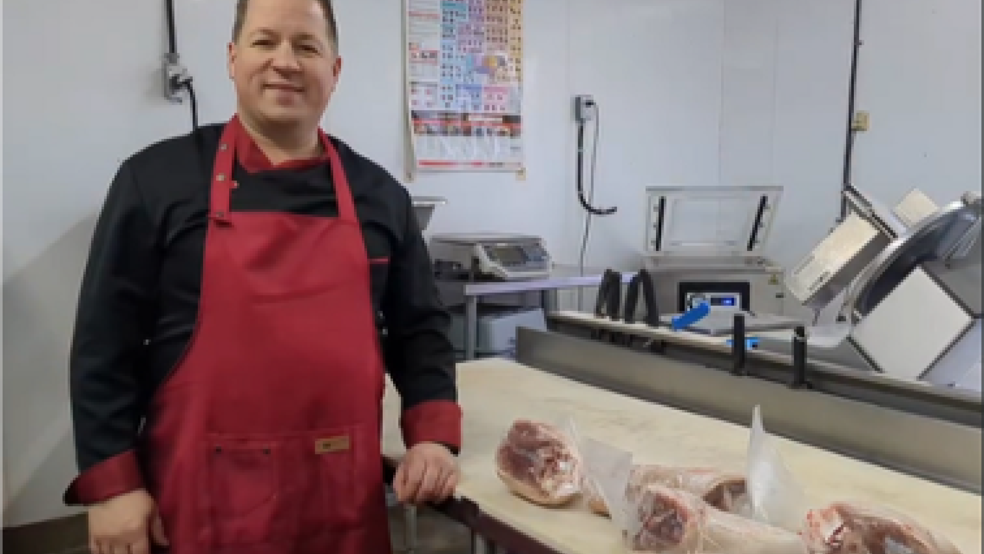 Butcher wearing red apron stands by a table with wrapped meat.