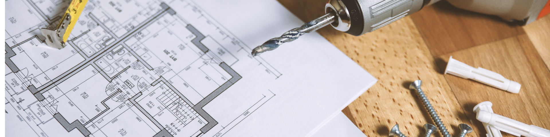 Blueprints on a table with a drill, screws, and a measuring tape.