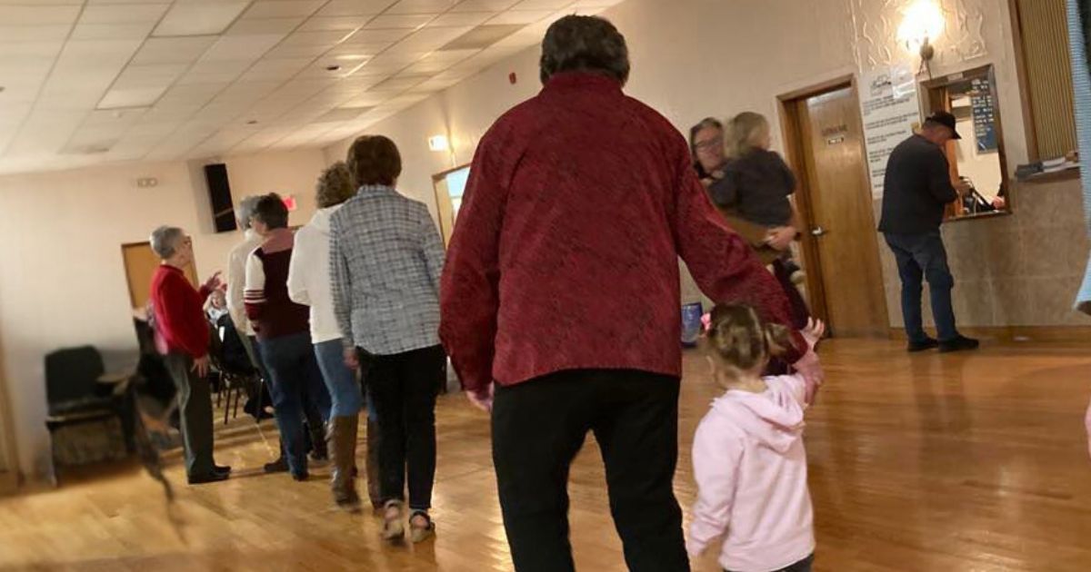 Older woman line dancing in community hall. A young girl is holding the older woman's hand. 